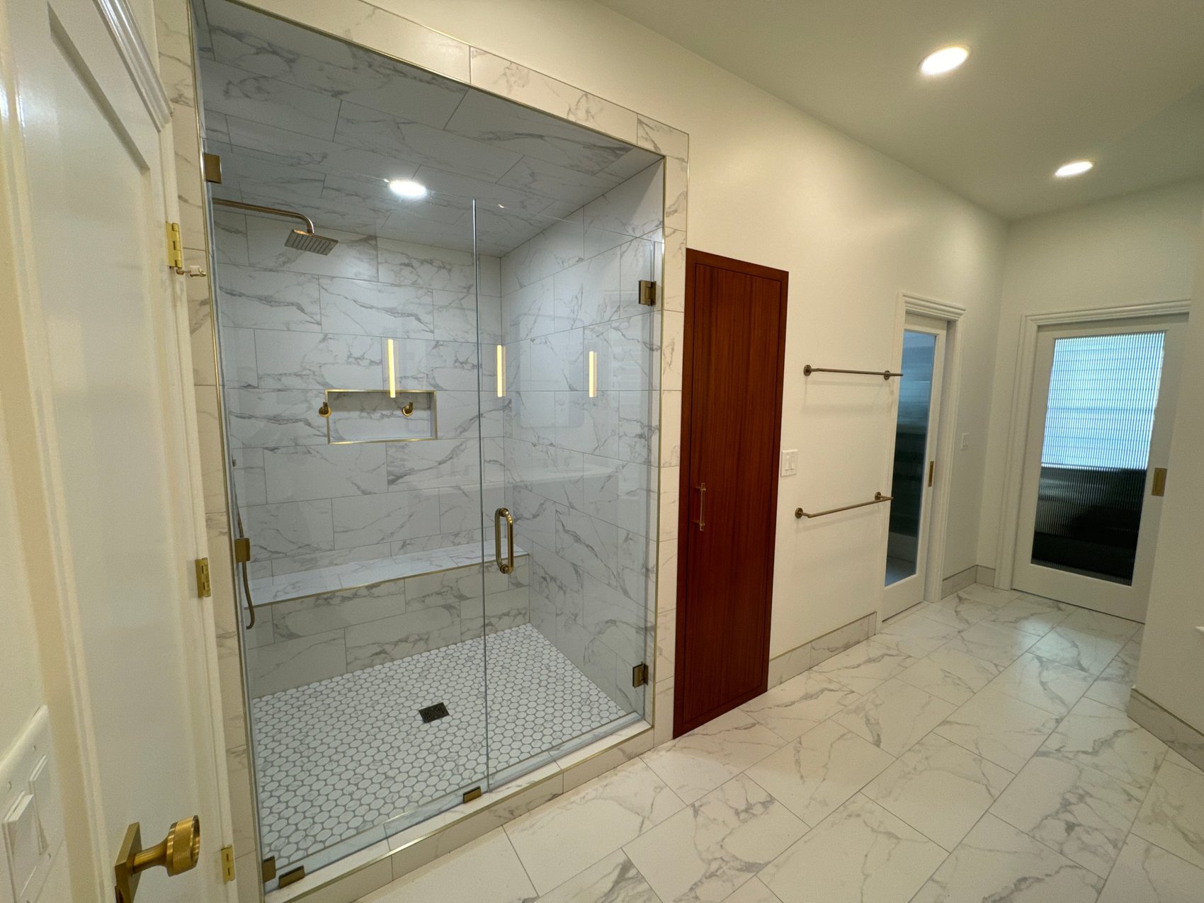 Remodeled bathroom with frameless glass shower, marble-look tile, bench and brass fixtures