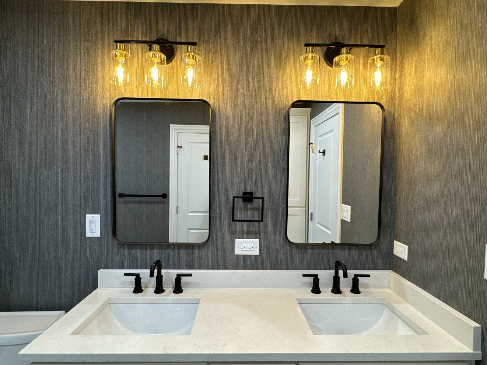 Bathroom with dark textured wallpaper, gold sconces, black mirrors and white double vanity